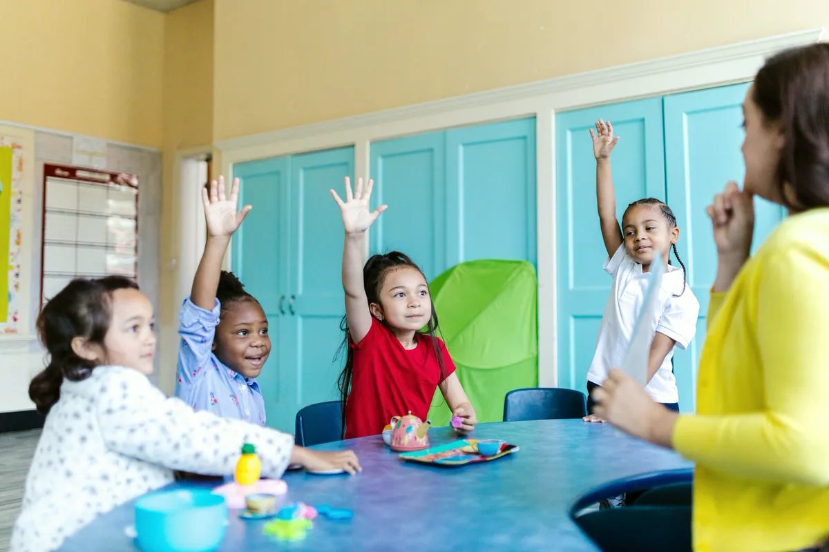 Young students eagerly raising their hands around a table in a bright classroom
