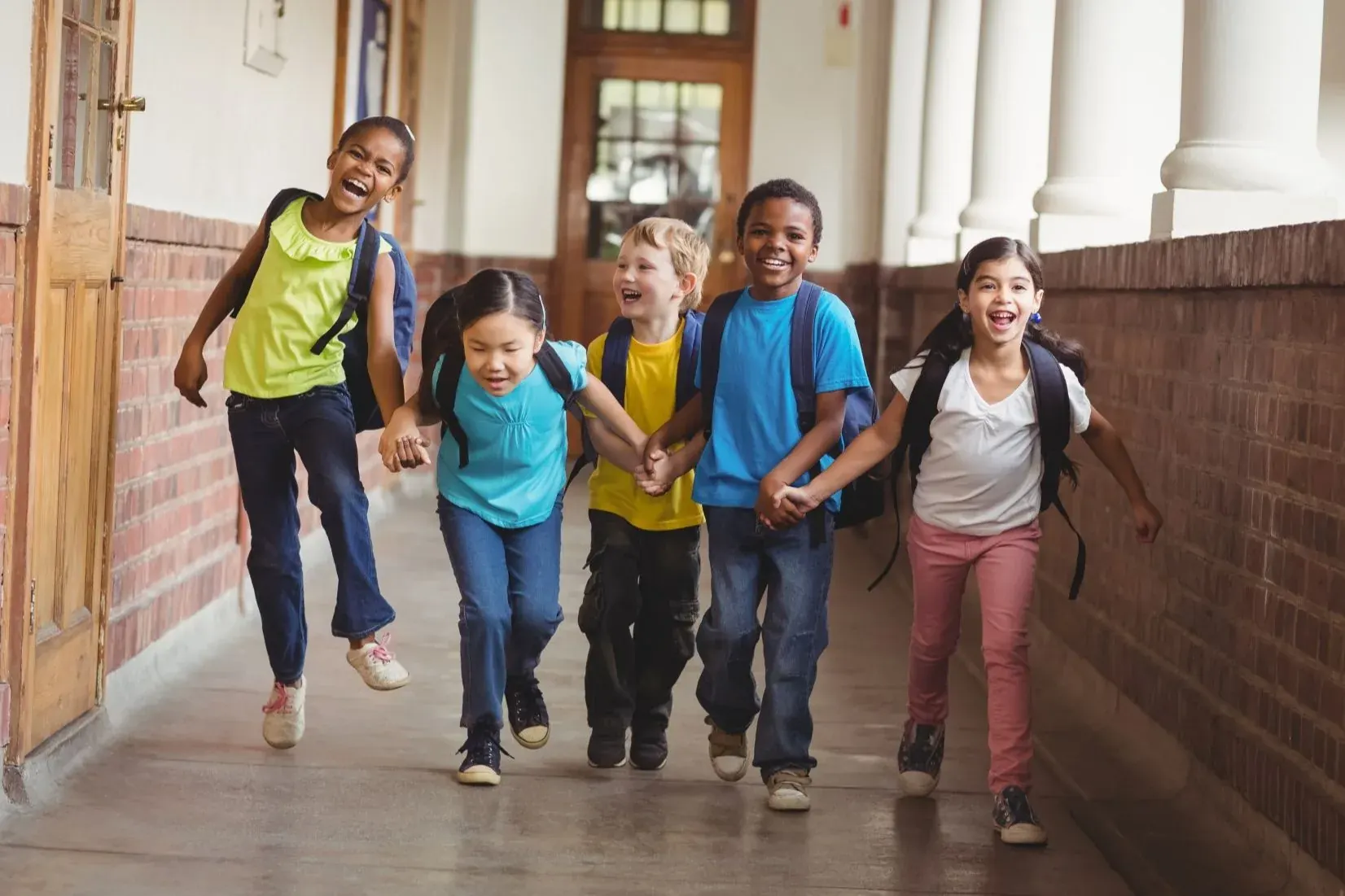 Five smiling children hold hands as they run down a school hallway
